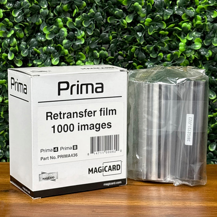 A box labeled "Magicard PRIMA436 Retransfer Film for the Magicard Prima 4 and Prima 8, 1,000 Prints" and a sealed roll of retransfer film rest on a wooden surface with a green leafy background.