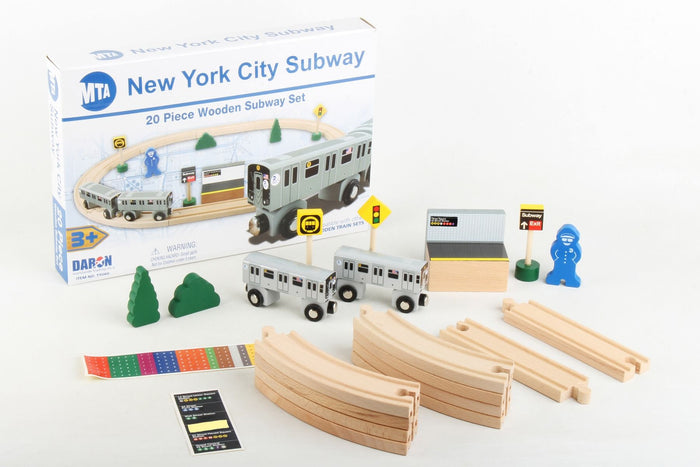 The image shows the packaging for the New York City Subway 20‑Piece Wooden Subway Set in the upper left. In front of it, the pieces are neatly arranged: two small green wooden trees, two silver‑gray subway cars, a wooden platform, three yellow signal signs—one with a traffic light, one with a train icon, and one with a subway‑exit symbol—a small wooden conductor dressed in blue, a stack of wooden track pieces at the bottom center, and a sheet of colorful decals for subway lines.
