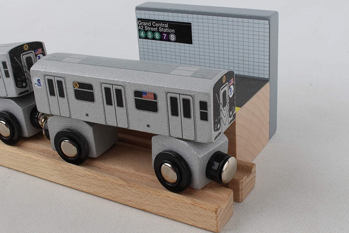 A silver‑gray MTA R‑train subway car sits on wooden tracks beside a black platform with a yellow edge. Behind it is a gray‑blue tiled wall with a sign reading “Grand Central – 42 Street Station,” along with the train line symbols.