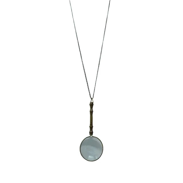 On a white background, the necklace displays an ornate magnifying glass pendant made of aged brass. The magnifying glass is about four inches long, with a decorative handle and a clear, working lens. It hangs from a matching aged brass scroll chain and closes with a small lobster clasp, giving the piece a refined, antique-inspired appearance.