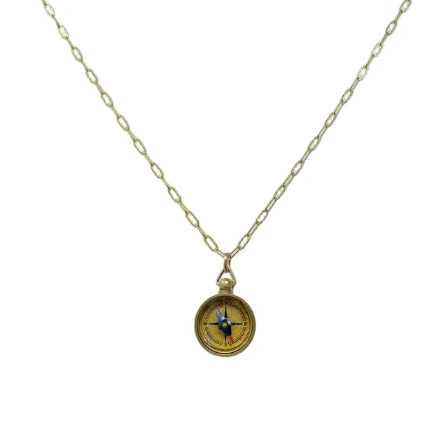 The image shows a delicate pendant necklace with a small, round compass charm about half an inch wide, finished in warm gold‑toned brass. The smooth compass face has lightly engraved directional markings and hangs from a slim, gold‑plated brass cable chain with a small lobster clasp.