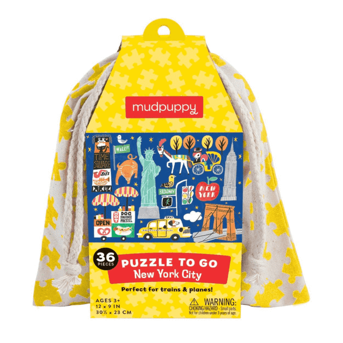 A bright yellow, house-shaped die-cut puzzle box titled “Mudpuppy: New York City” featuring high-contrast illustrations of the Statue of Liberty, Brooklyn Bridge, and a striped hot dog stand. A yellow handle at the roof peak allows a drawstring bag to flow down the sides, with product details printed along the bottom left and right edges.