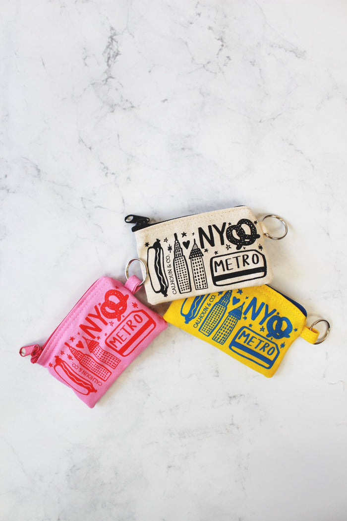 New York Screen Printed Coin Pouch, available in pink, white, and yellow: A small, soft fabric pouch with a smooth, flat surface and a zipper along the top edge. The pouch is rectangular and lightweight, easy to hold in one hand. The front is printed with bold, high‑contrast New York–themed graphics—clean shapes and simple lettering—with no raised textures. The fabric feels flexible but sturdy, designed to hold coins or small items securely.