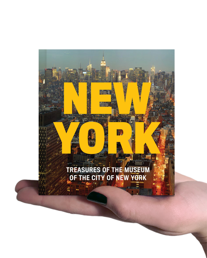 Pocket-sized book held in an open palm, showing the cover of New York: Treasures from the Museum of the City of New York. The cover features the title "New York" in large, bold yellow lettering above the subtitle, set against a vibrant photograph of the New York City skyline at dusk, glowing with colorful city lights beneath a deep blue evening sky.