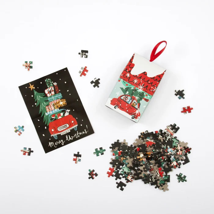 A finished puzzle shows a red car overflowing with gifts and a green Christmas tree against a black background. To the right sits a red‑and‑green, house‑shaped cardboard ornament that also holds the puzzle, illustrated with the same cheerful gift‑packed car inside a cozy holiday house. Below, the puzzle is shown unassembled, with a few loose pieces scattered all around.