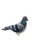 A small pigeon brooch with a simple, elongated body and rounded head. Its surface is richly textured with soft fabric, raised stitching, and beadwork, with sequins adding a gentle shimmer in shades of green, grey, and black. A smooth metal pin is attached to the back for fastening, and each handmade piece features slight variations in texture and detail.