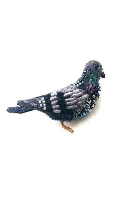 A small pigeon brooch with a simple, elongated body and rounded head. Its surface is richly textured with soft fabric, raised stitching, and beadwork, with sequins adding a gentle shimmer in shades of green, grey, and black. A smooth metal pin is attached to the back for fastening, and each handmade piece features slight variations in texture and detail.