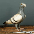 Features a solid cast iron pigeon figurine, hand-painted in soft shades of grayish blue, white, and black, with a lightly distressed finish that gives it a vintage look; each piece is individually hand-finished, so slight variations in color and detailing are to be expected. The figurine is displayed resting on a wooden table, set against a green wall backdrop.