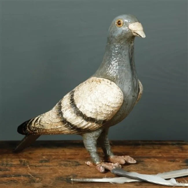 Features a solid cast iron pigeon figurine, hand-painted in soft shades of grayish blue, white, and black, with a lightly distressed finish that gives it a vintage look; each piece is individually hand-finished, so slight variations in color and detailing are to be expected. The figurine is displayed resting on a wooden table, set against a green wall backdrop.