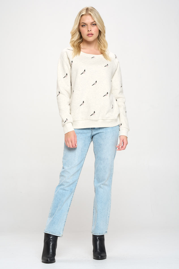 Image shows a full view of a woman wearing a crew neck heather gray sweatshirt with a pigeon print evenly repeated across the front, sleeves, and back, forming a continuous all‑over pattern rather than a single central graphic.
