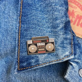 An enamel pin shaped like a 1980s boombox, attached to the collar of a denim jacket. The pin is mostly black with gold and red details. It features two large, round speakers on either side and cassette player buttons and controls in the center.