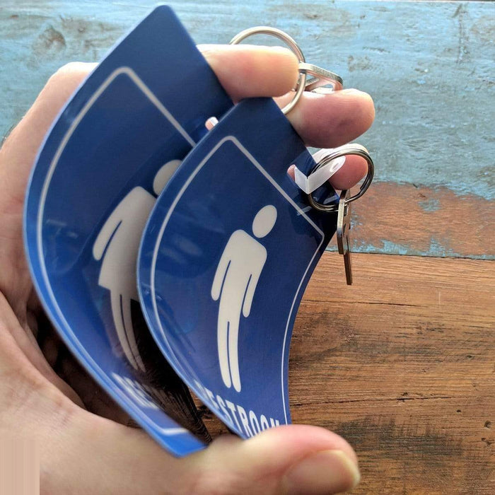 Hand holding two bent blue Restroom Pass Keychain - Bathroom Tag with Key Chain Ring - Heavy Duty Large Passes for Men and Women Restrooms with Key Holder (Sold in 2 Pack) (SPID-9830-Blue); each pass is attached to a bathroom keychain. Background shows a wooden surface.