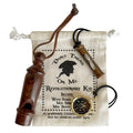 The image shows a Revolutionary War–themed kit on a white background. On top of a muslin drawstring bag printed with “Don’t Tread on Me” are a small wooden whistle, a 2‑inch brass sand timer necklace, and a gold‑colored compass with a black dial. The materials—wood, metal, and soft fabric—are easy to distinguish by touch.