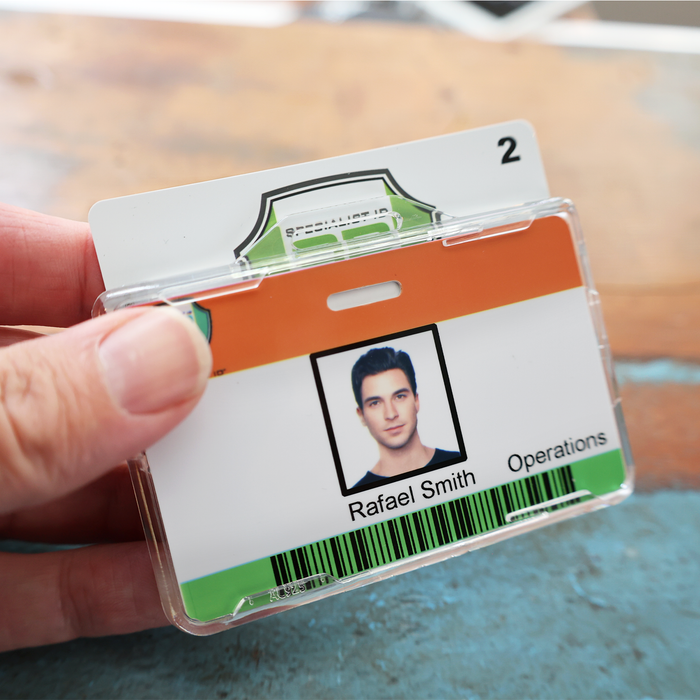 A hand holds an employee ID badge for "Rafael Smith, Operations" in a Specialist ID Dual Sided RFID Blocking Badge Holder (SPID-1760), a heavy duty protector for secure badges, featuring a photo, barcode, and the number 2 at the top.