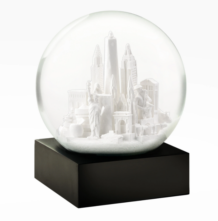 Snow Globe: NYC White — 
A round glass snow globe about 4 inches wide and 4.5 inches tall. Inside the clear globe is a small white model of New York City landmarks, including simplified shapes of the Chrysler Building, One World Trade Center, and the Wall Street bull. The buildings are smooth with no raised texture. The globe sits on a solid square black base with flat sides. When shaken, fine white flakes swirl around the miniature skyline. The overall shape is compact.