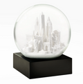 Snow Globe: NYC White — 
A round glass snow globe about 4 inches wide and 4.5 inches tall. Inside the clear globe is a small white model of New York City landmarks, including simplified shapes of the Chrysler Building, One World Trade Center, and the Wall Street bull. The buildings are smooth with no raised texture. The globe sits on a solid square black base with flat sides. When shaken, fine white flakes swirl around the miniature skyline. The overall shape is compact.