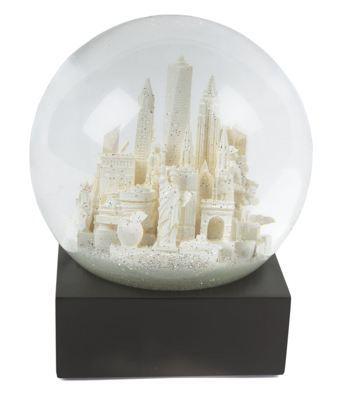 Snow Globe: NYC White — 
A round glass snow globe about 4 inches wide and 4.5 inches tall. Inside the clear globe is a small white model of New York City landmarks, including simplified shapes of the Chrysler Building, One World Trade Center, and the Wall Street bull. The buildings are smooth with no raised texture. The globe sits on a solid square black base with flat sides. When shaken, fine white flakes swirl around the miniature skyline. The overall shape is compact.