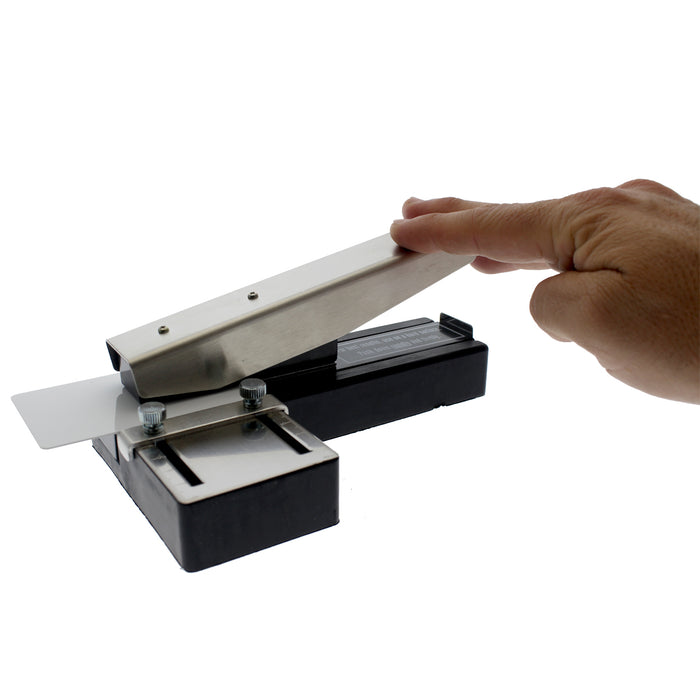 A hand presses down on a metal and plastic device with a small rectangular opening, similar to an ID badge slot punch. This **Stapler-Style Slot Punch With Adjustable Guide (P/N SPID-9750)** features a non-slip rubber base for stable operation.