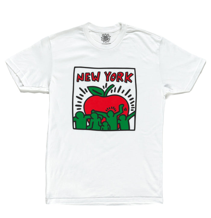 A soft white T shirt featuring a large, smooth graphic of a stylized apple in Keith Haring’s signature bold style. The apple is outlined with thick black lines and filled with lively red and green human figures that create a playful, energetic feel against the white background. The shirt has a classic unisex fit with short sleeves and a standard crew neckline. It’s lightweight, comfortable, and available in sizes from small to extra large.