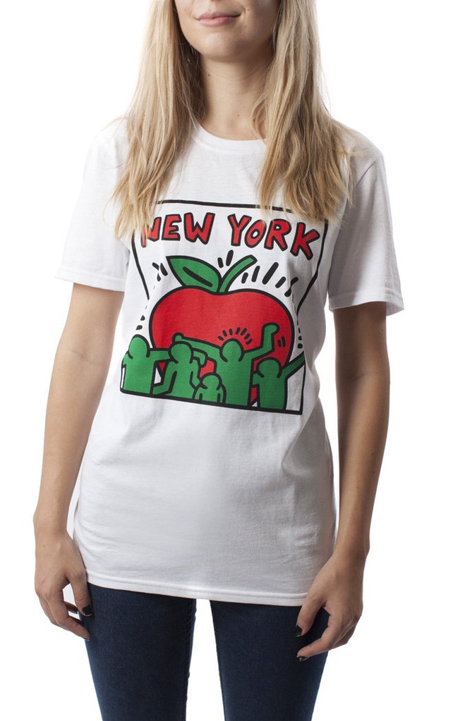 A woman wears a soft white T‑shirt with a large Keith Haring–style apple graphic on the front. The apple has thick black outlines and is filled with bright red and green dancing figures. The shirt has short sleeves, a crew neck, and a classic unisex fit. It’s lightweight, comfortable, and available in sizes from small to extra large.