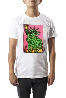 A boy is wearing a white T‑shirt featuring a bold black‑outlined image of a green Statue of Liberty holding a yellow flame. The figure stands on a pink background, with playful yellow human figures in the foreground along the bottom edge of the rectangular Keith Haring design.