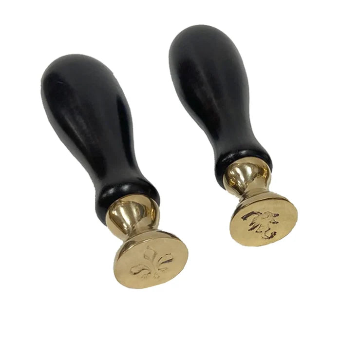 Two antique‑style wax seal stamps—a fleur‑de‑lis and a leaf—shown on a white background. Each has a dark cherry‑stained wooden handle and a 1⅛‑inch round solid brass seal with a distressed finish. The seals are about 5½ inches tall, interchangeable, and designed for use with traditional hard sealing wax. Sold separately.