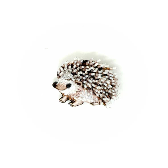 White Breasted Hedgehog Brooch - A small, rounded hedgehog brooch with a compact body and slightly pointed snout. The surface mixes soft felt with raised stitching, beads, and sequins to suggest textured spines and a fluffy chest. It has a layered, tactile feel with subtle sparkle, and a smooth metal pin on the back for fastening. Each handmade piece varies slightly in texture and detail.