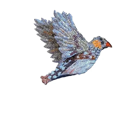 A close-up of a hand‑embroidered Zebra Finch brooch on a plain background. The small bird is shown in profile, mid‑flight with its wings outstretched, featuring a soft light blue body, a bright orange cheek patch, a spotted tail, and a vivid red beak. The surface appears textured and slightly raised, crafted from beads, sequins, stones, yarns, and fine fabrics, giving it a richly detailed, tactile quality.